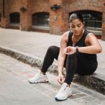 Mulher usando roupa esportiva verifica um smartwatch enquanto está sentada em uma calçada urbana.
