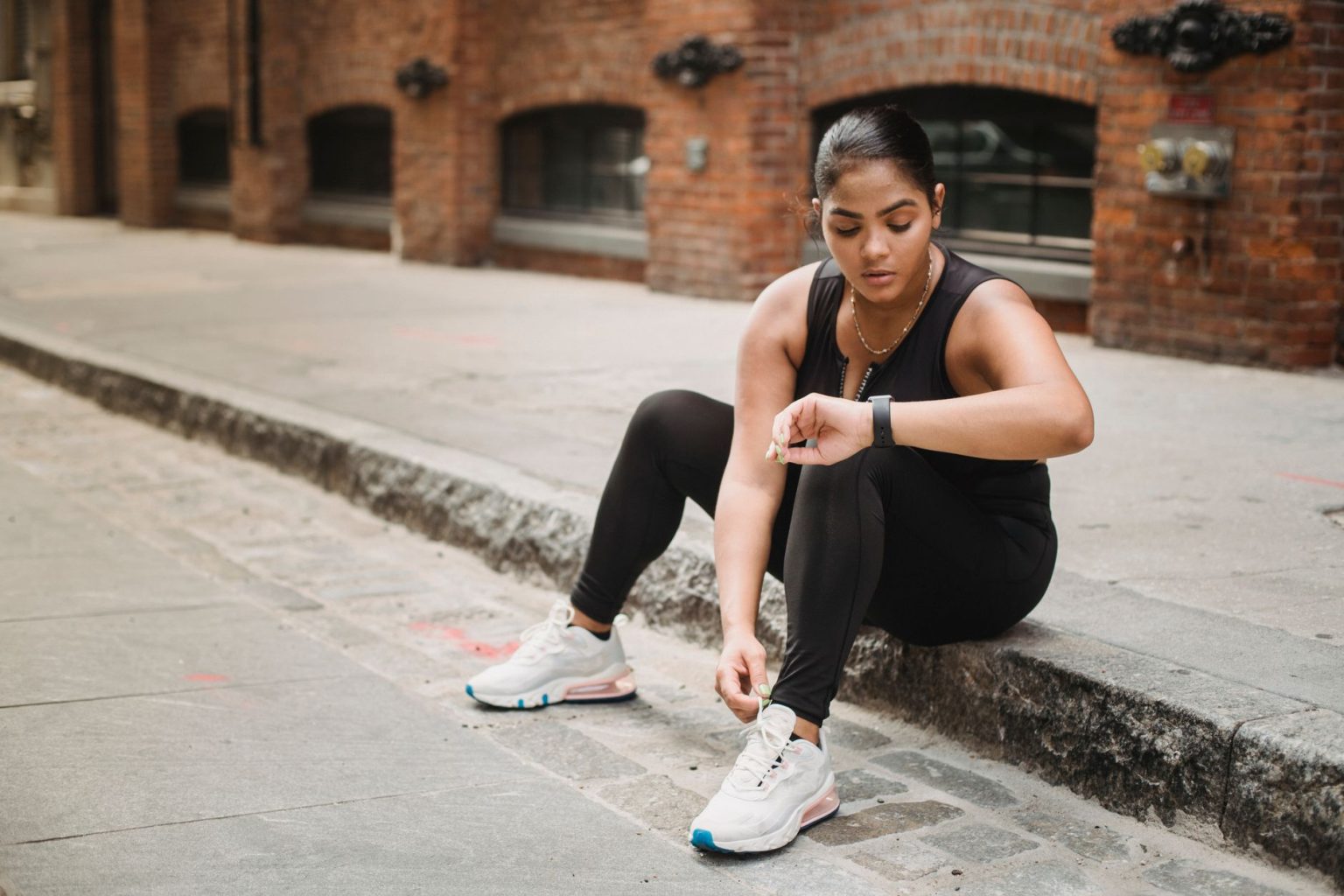 Mulher usando roupa esportiva verifica um smartwatch enquanto está sentada em uma calçada urbana.