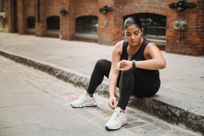 Mulher usando roupa esportiva verifica um smartwatch enquanto está sentada em uma calçada urbana.