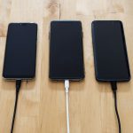 Três smartphones conectados a carregadores sobre uma superfície de madeira.