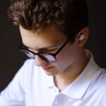 Jovem usando óculos e fones de ouvido, concentrado enquanto escuta áudio em um ambiente interno.
