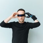 Homem ajusta óculos escuros de aparência futurista diante de um fundo neutro.