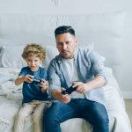 Pai acompanha o filho enquanto os dois jogam videogame sentados em uma cama