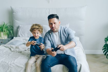 Pai acompanha o filho enquanto os dois jogam videogame sentados em uma cama