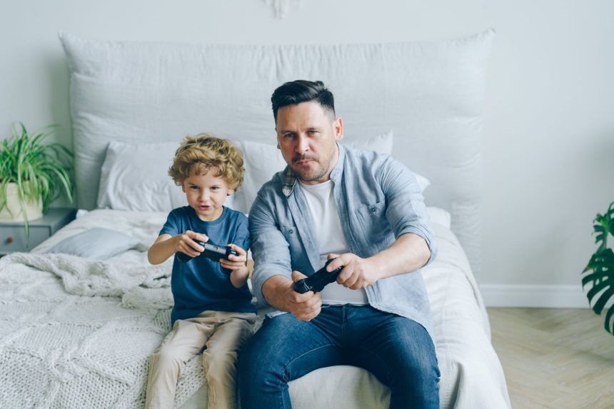 Pai acompanha o filho enquanto os dois jogam videogame sentados em uma cama